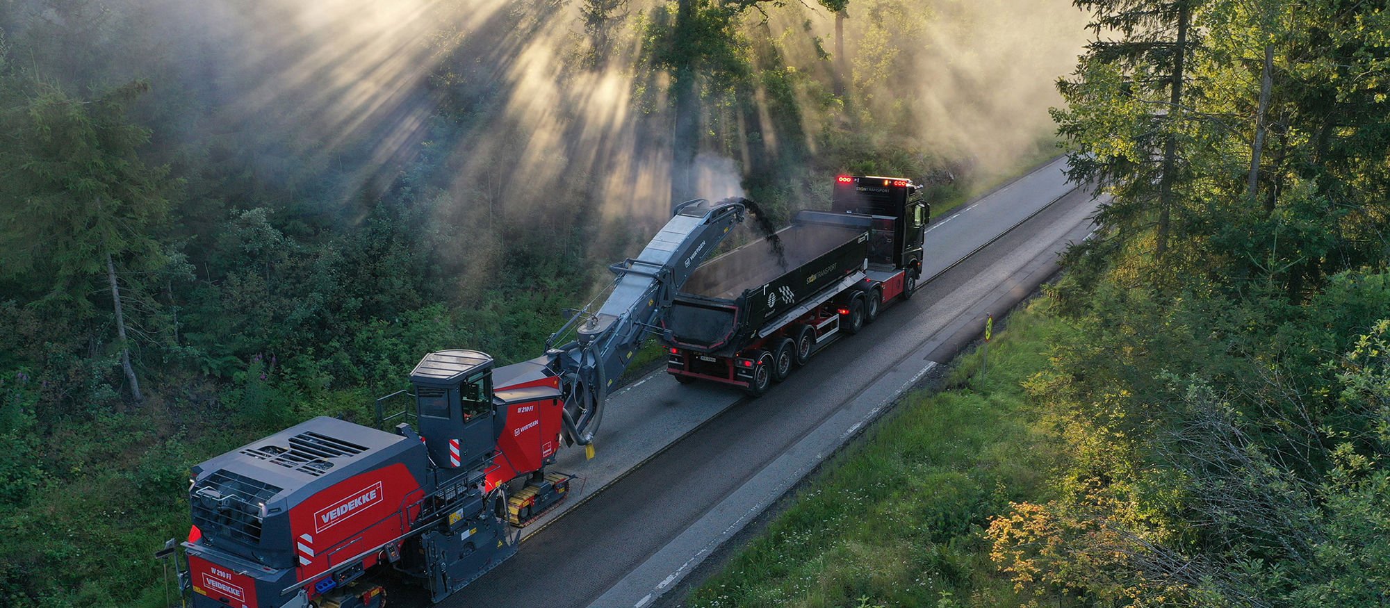 Asfaltfres og lastebil på vei med solstråler gjennom skog. Foto.